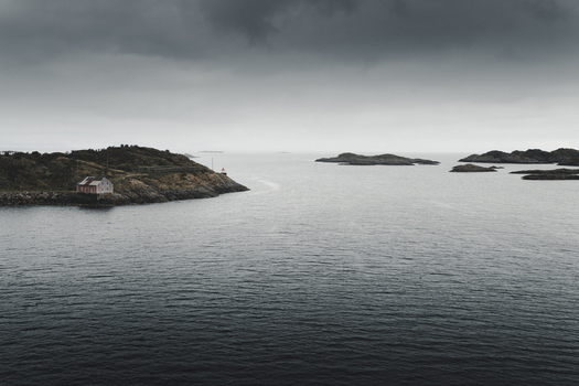 View of a traditional red wooden house on the rocky coastline with calm dark sea and distant islands under an overcast sky in Henningsvaer Nordland Norway. View of a traditional red wooden house on the rocky coastline with calm dark sea and distant islands under an overcast sky in Henningsvaer Nordland Norway.