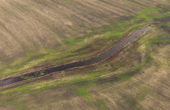 Aerial view of a winding stream cutting through agricultural fields with patches of green grass and brown soil in Pomeranian Voivodeship Poland.