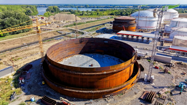 Aerial view of the assembly of a large metal oil storage tank at an industrial refinery site with a construction crane surrounding infrastructure and a river in Kovin Vojvodina Serbia. Aerial view of the assembly of a large metal oil storage tank at an industrial refinery site with a construction crane surrounding infrastructure and a river in Kovin Vojvodina Serbia.