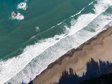 Aerial view of the southern Oregon coast showing waves crashing onto the sandy beach with dark rock formations in the turquoise water and tree shadows in Gold Beach Oregon United States. Aerial view of the southern Oregon coast showing waves crashing onto the sandy beach with dark rock formations in the turquoise water and tree shadows in Gold Beach Oregon United States.