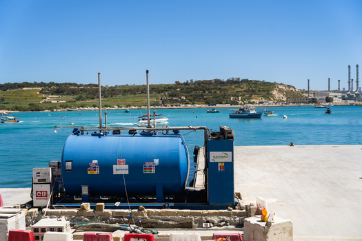 View of a blue fuel tank on a concrete pier overlooking the turquoise waters of the bay with boats and industrial chimneys in the background Marsaskala Southern Region Malta. View of a blue fuel tank on a concrete pier overlooking the turquoise waters of the bay with boats and industrial chimneys in the background Marsaskala Southern Region Malta.