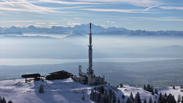 Aerial view of a stark white telecommunications tower piercing through the mist shrouded valley with snow dusted peaks in the distance Gex Auvergne Rhône Alpes France. Aerial view of a stark white telecommunications tower piercing through the mist shrouded valley with snow dusted peaks in the distance Gex Auvergne Rhône Alpes France.
