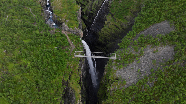 Aerial view of Gorsa bridge spanning a deep canyon with a powerful waterfall surrounded by lush green forests and rugged rocky terrain in Birtavarre Troms Norway. Aerial view of Gorsa bridge spanning a deep canyon with a powerful waterfall surrounded by lush green forests and rugged rocky terrain in Birtavarre Troms Norway.