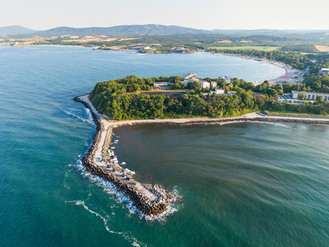 Aerial view of the coastal landscape featuring a long breakwater extending into the Black Sea surrounded by lush green trees sandy beaches and calm blue waters in Kiten Burgas Bulgaria. Aerial view of the coastal landscape featuring a long breakwater extending into the Black Sea surrounded by lush green trees sandy beaches and calm blue waters in Kiten Burgas Bulgaria.