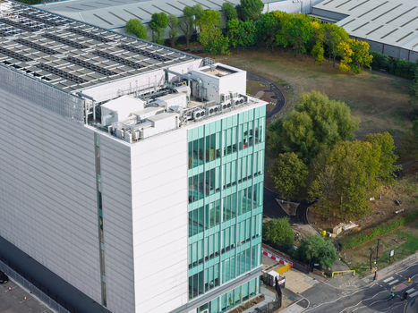 Aerial view of a modern data center with a glass facade and rooftop cooling units surrounded by green trees and a road in the United Kingdom. Aerial view of a modern data center with a glass facade and rooftop cooling units surrounded by green trees and a road in the United Kingdom.