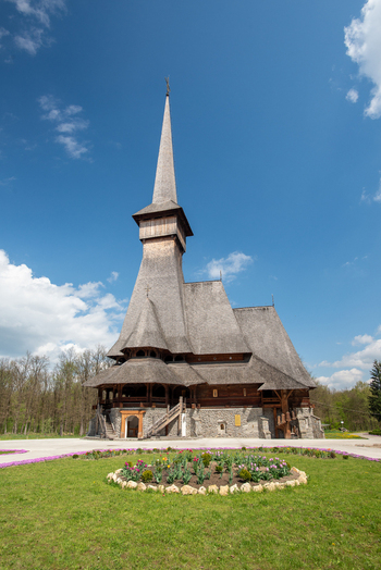 View of the traditional wooden church with its tall spire shingled roof and stone foundation set against a clear blue sky in Maramures Romania. View of the traditional wooden church with its tall spire shingled roof and stone foundation set against a clear blue sky in Maramures Romania.