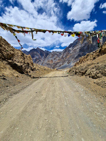 View of the rugged Penzi La road in Zanskar Ladakh India featuring colorful prayer flags strung across the dirt path against a backdrop of dramatic mountain peaks and a blue sky.