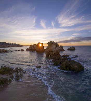 Aerial view of Praia dos Arrifes featuring dramatic rock formations in the ocean at sunset with the sun shining through a natural arch in the rock sandy beach in Faro District Portugal. Aerial view of Praia dos Arrifes featuring dramatic rock formations in the ocean at sunset with the sun shining through a natural arch in the rock sandy beach in Faro District Portugal.
