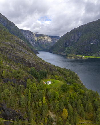 Aerial view of a small white house and a red shed nestled on a lush green hillside overlooking a calm fjord surrounded by steep rocky mountains under a cloudy sky Fjaera Vestland Norway. Aerial view of a small white house and a red shed nestled on a lush green hillside overlooking a calm fjord surrounded by steep rocky mountains under a cloudy sky Fjaera Vestland Norway.