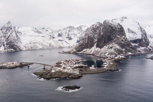 Aerial view of Lofoten featuring a small coastal village connected by a bridge surrounded by snow covered mountains and dark sea in Nordland Norway.