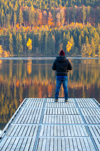 View of a man standing on a wooden pier overlooking a calm lake with reflections of autumn forest in Breitnau Baden Wurttemberg Germany. View of a man standing on a wooden pier overlooking a calm lake with reflections of autumn forest in Breitnau Baden Wurttemberg Germany.