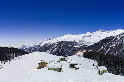 View of snow covered stone huts on a mountain slope under a starry night sky with distant alpine peaks Madesimo Lombardy Italy. View of snow covered stone huts on a mountain slope under a starry night sky with distant alpine peaks Madesimo Lombardy Italy.