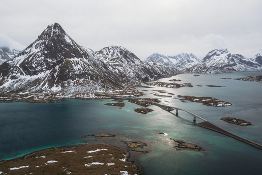 Aerial view of the bridge stretching across the turquoise waters connecting the snow dusted isles beneath the towering jagged peaks Flakstadbruene Nordland Norway. Aerial view of the bridge stretching across the turquoise waters connecting the snow dusted isles beneath the towering jagged peaks Flakstadbruene Nordland Norway.