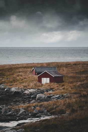 View of red wooden cabins situated in a coastal landscape with dry grass rocky terrain and a dramatic cloudy sky in Ramberg Nordland Norway. View of red wooden cabins situated in a coastal landscape with dry grass rocky terrain and a dramatic cloudy sky in Ramberg Nordland Norway.