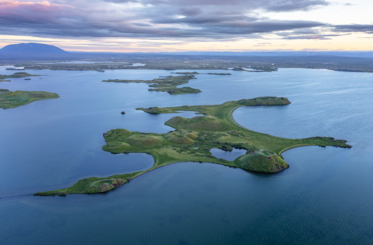 Aerial view of Lake Myvatn featuring volcanic craters and lush green islands surrounded by calm blue water under a soft sunset sky in Thingeyjarsveit Iceland.