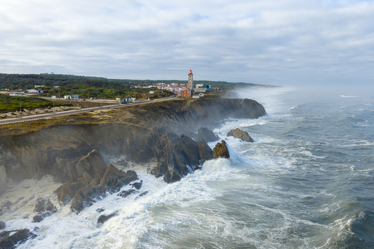 Aerial view of Farol Penedo da Saudade lighthouse standing on rugged cliffs with crashing waves and coastal mist Marinha Grande Leiria Portugal. Aerial view of Farol Penedo da Saudade lighthouse standing on rugged cliffs with crashing waves and coastal mist Marinha Grande Leiria Portugal.