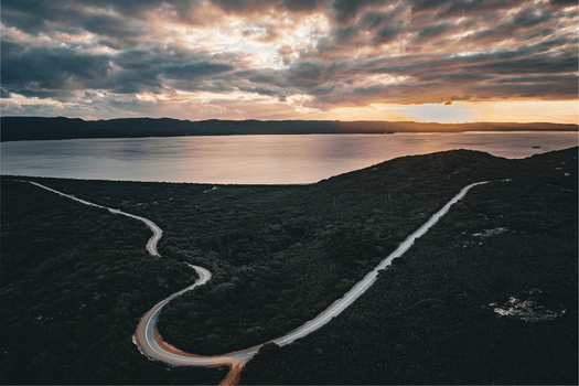 Aerial view of Torndirrup National Park featuring a winding road through dense coastal vegetation overlooking the bay under a dramatic sunset sky in Albany Western Australia Australia. Aerial view of Torndirrup National Park featuring a winding road through dense coastal vegetation overlooking the bay under a dramatic sunset sky in Albany Western Australia Australia.