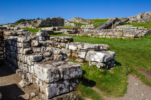 View of the ancient Roman ruins of Vindolanda featuring stone walls and grassy foundations under a clear blue sky in Hexham England United Kingdom. View of the ancient Roman ruins of Vindolanda featuring stone walls and grassy foundations under a clear blue sky in Hexham England United Kingdom.