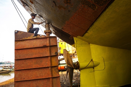 Dhaka Bangladesh 20 April 2026 View of a worker painting a ship s hull contrasting vibrant yellow against dark weathered rust and industrial machinery.