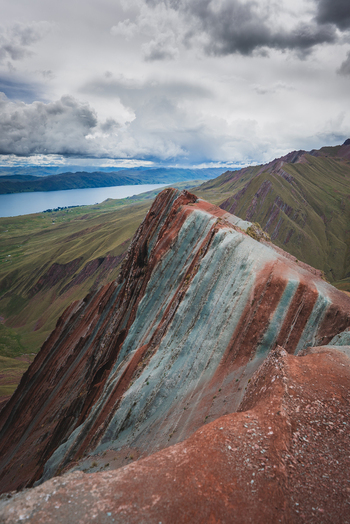 View of the vibrant multi colored mineral layers of Pallay Punchu Rainbow Mountain overlooking a vast lake under a dramatic cloudy sky in Layo Cuzco Peru. View of the vibrant multi colored mineral layers of Pallay Punchu Rainbow Mountain overlooking a vast lake under a dramatic cloudy sky in Layo Cuzco Peru.