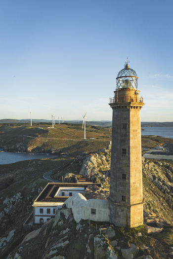 Aerial view of the historic Faro de Cabo Vilan lighthouse perched on rocky cliffs with wind turbines in the background under a clear blue sky in Galicia Spain.