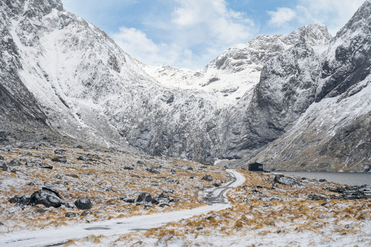 View of a dirt path leading to a small cabin by a lake surrounded by snow covered mountains in Leknes Lofoten Norway.