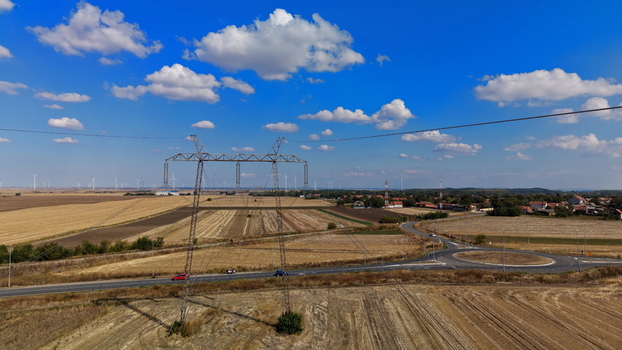 Aerial view of electric power transmission lines spanning over rural fields a roundabout and a road with wind turbines in the distance in Pancevo Serbia.