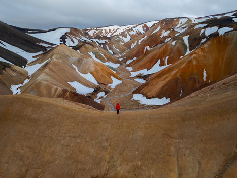 Aerial view of Landmannalaugar with a person in a red jacket on a ridge overlooking colorful rhyolite mountains with patches of snow and a winding stream in Rangarthing ytra Iceland. Aerial view of Landmannalaugar with a person in a red jacket on a ridge overlooking colorful rhyolite mountains with patches of snow and a winding stream in Rangarthing ytra Iceland.