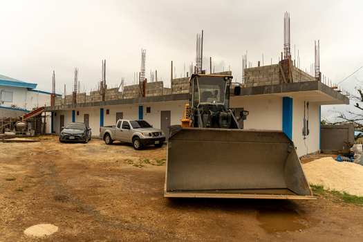 San Jose Tinian Northern Mariana Islands 05 July 2026 View of a bulldozer parked at a construction site with an unfinished building featuring exposed rebar and parked cars under an overcast sky. San Jose Tinian Northern Mariana Islands 05 July 2026 View of a bulldozer parked at a construction site with an unfinished building featuring exposed rebar and parked cars under an overcast sky.