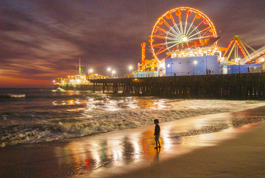 View of the illuminated Ferris wheel on Santa Monica Pier at night with a child standing on the sandy beach and gentle waves rolling in Santa Monica California United States. View of the illuminated Ferris wheel on Santa Monica Pier at night with a child standing on the sandy beach and gentle waves rolling in Santa Monica California United States.