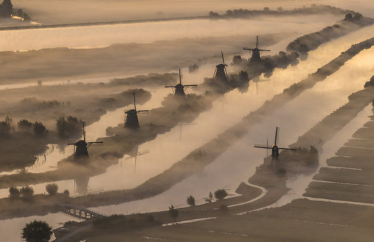 Aerial view of historic windmills shrouded in thick morning mist along the canal during a golden sunrise Kinderdijk Zuid Holland Netherlands.