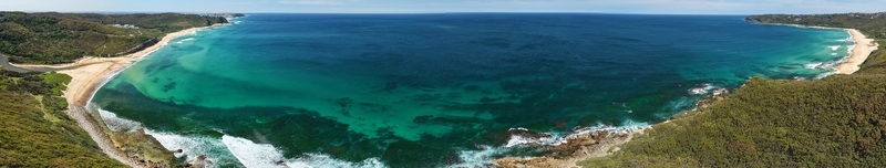 Aerial view of turquoise waters meet sandy shores embraced by lush green headlands under a clear sky creating a vibrant coastal panorama Newcastle New South Wales Australia.