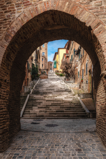 View of the historic Piaggia staircase framed by a brick archway with old stone buildings and a clear blue sky in Corinaldo Marche Italy. View of the historic Piaggia staircase framed by a brick archway with old stone buildings and a clear blue sky in Corinaldo Marche Italy.