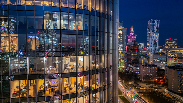 Aerial view of the illuminated glass facade of a modern skyscraper with office interiors and the Warsaw city skyline featuring the Palace of Culture and Science at night in Poland. Aerial view of the illuminated glass facade of a modern skyscraper with office interiors and the Warsaw city skyline featuring the Palace of Culture and Science at night in Poland.