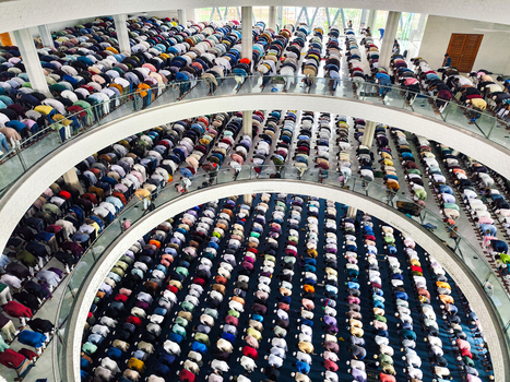Dhaka Bangladesh 19 June 2026 Aerial view of a large crowd of men praying in rows inside a modern multi level mosque with curved balconies and bright interior lighting.
