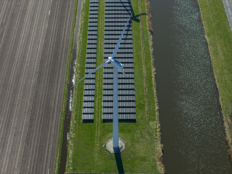 Aerial view of a tall white wind turbine and rows of solar panels situated between a canal and a plowed field under bright sunlight Netherlands. Aerial view of a tall white wind turbine and rows of solar panels situated between a canal and a plowed field under bright sunlight Netherlands.
