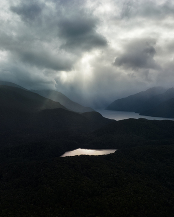 Aerial view of sun rays piercing through stormy clouds over the rugged shadowed landscape illuminating a small lake near Manapouri Lake Manapouri Southland Region New Zealand. Aerial view of sun rays piercing through stormy clouds over the rugged shadowed landscape illuminating a small lake near Manapouri Lake Manapouri Southland Region New Zealand.