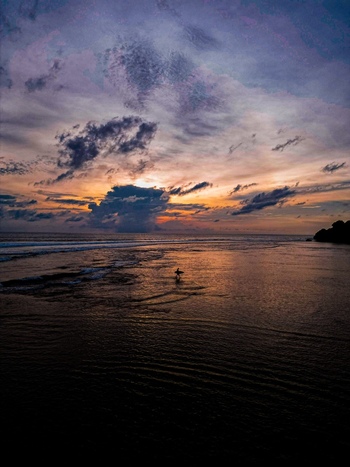 Aerial view of Pantai Nunggalan at sunset with a lone surfer walking in the shallow water calm waves and dramatic clouds in Kecamatan Kuta Selatan Bali Indonesia.