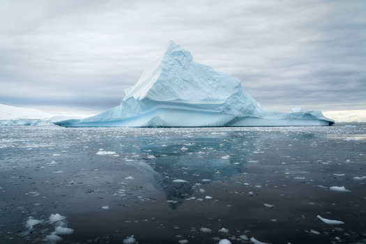 View of a massive pointed iceberg floating in the calm dark waters with smaller ice fragments and a reflection under an overcast sky in Antarctica.