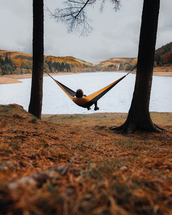 Hope Valley England United Kingdom 03 November 2021 View of a person relaxing in a hammock suspended between two trees overlooking the calm waters of Ladybower Reservoir during autumn.