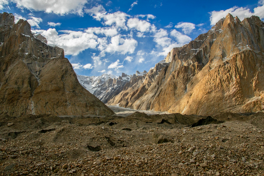 View of the granite spires of the Trango Towers rising above a rocky glacial moraine under a bright blue sky with scattered clouds in Pakistan. View of the granite spires of the Trango Towers rising above a rocky glacial moraine under a bright blue sky with scattered clouds in Pakistan.