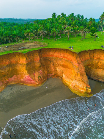 Aerial view of dramatic orange cliffs overlooking a sandy beach with lush palm trees and people walking along the shore in Bali Bali Indonesia. Aerial view of dramatic orange cliffs overlooking a sandy beach with lush palm trees and people walking along the shore in Bali Bali Indonesia.