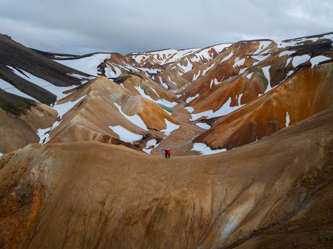 Aerial view of Landmannalaugar with two people standing on a ridge overlooking colorful rhyolite mountains snow patches and a winding stream in Rangarthing ytra Iceland. Aerial view of Landmannalaugar with two people standing on a ridge overlooking colorful rhyolite mountains snow patches and a winding stream in Rangarthing ytra Iceland.