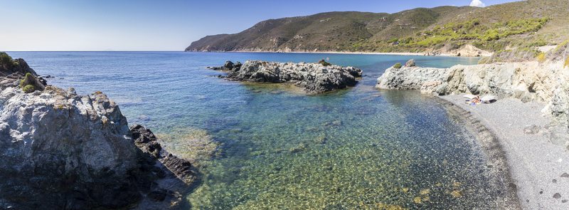 View of clear turquoise waters meeting rugged rocky shores leading to a pebbled beach under a bright sky in Lacona Elba Italy. View of clear turquoise waters meeting rugged rocky shores leading to a pebbled beach under a bright sky in Lacona Elba Italy.