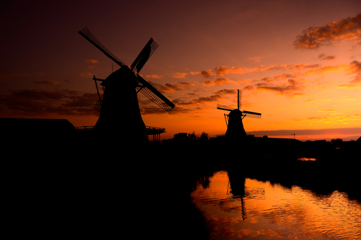 View of two traditional windmills silhouetted against a vibrant orange sunset with reflections in a calm canal in the Netherlands. View of two traditional windmills silhouetted against a vibrant orange sunset with reflections in a calm canal in the Netherlands.