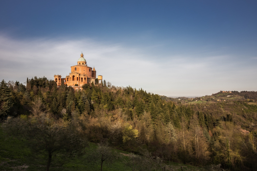 View of Santuario Madonna di San Luca perched on a lush green hill surrounded by dense forest under a clear blue sky Bologna Emilia Romagna Italy.