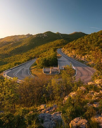Aerial view of a winding mountain road with a sharp hairpin turn and a stone monument surrounded by lush green hills during golden hour in Rijeka Primorje Gorski Kotar Croatia. Aerial view of a winding mountain road with a sharp hairpin turn and a stone monument surrounded by lush green hills during golden hour in Rijeka Primorje Gorski Kotar Croatia.