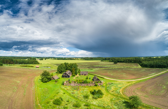 Aerial view of a rural farmstead with stone walls and wooden buildings surrounded by agricultural fields and lush green trees under a dramatic cloudy sky in Latvia. Aerial view of a rural farmstead with stone walls and wooden buildings surrounded by agricultural fields and lush green trees under a dramatic cloudy sky in Latvia.