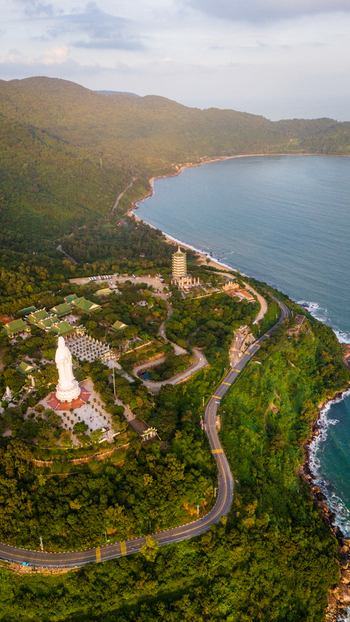Aerial view of Linh Ung Pagoda with its iconic giant Lady Buddha statue surrounded by lush green hills and a winding coastal road overlooking the sea in Da Nang Vietnam. Aerial view of Linh Ung Pagoda with its iconic giant Lady Buddha statue surrounded by lush green hills and a winding coastal road overlooking the sea in Da Nang Vietnam.
