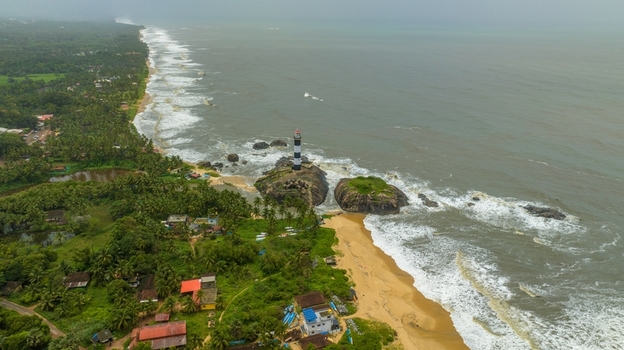 Aerial view of Kapu Light House standing on rocky outcrops along the coastline with lush palm trees sandy beach and crashing waves in Udupi Karnataka India.